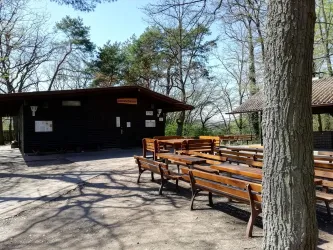 Ein rustikales Holzgebäude steht inmitten eines bewaldeten Bereichs. Rund um das Gebäude sind mehrere Holztische und Bänke aufgestellt.