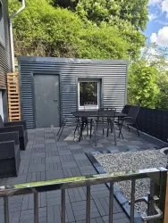 Eine gemütliche Terrasse mit einem Tisch und Stühlen aus Metall. Der Bereich ist modern gestaltet und von grünen Bäumen umgeben.