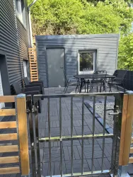 Eine moderne Terrasse mit einem Tisch und Stühlen. Im Hintergrund steht ein kleiner grauer Anbau umgeben von Bäumen.