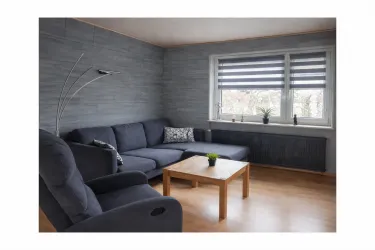 Ein modernes Wohnzimmer mit einer blauen Wohnlandschaft und einem Holztisch. Große Fenster ermöglichen viel Tageslicht und kleine Pflanzen verleihen dem Raum eine freundliche Atmosphäre.