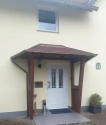 Ein Haus mit einer hellgelben Fassade und einem überdachten Eingangsbereich. Die Eingangstür ist weiß und ein kleiner Pflanzkübel steht neben der Treppe.