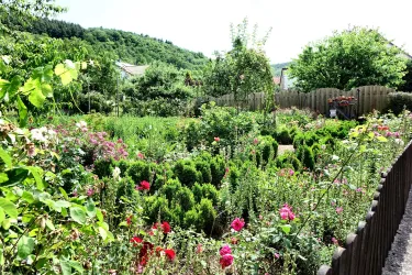 Ein blühender Garten mit vielen bunten Blumen und grünen Sträuchern. Im Hintergrund sind sanfte Hügel und ein Holzzaun zu sehen.