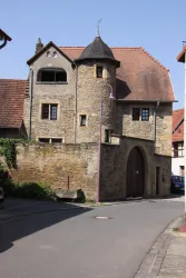 Ein historisches Gebäude mit einer steinernen Fassade und einem charakteristischen Dach. Die Straße vor dem Gebäude ist gepflastert und führt um die Ecke.
