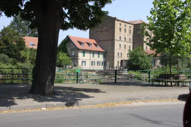 Eine alte Fabrik mit großen Fenstern und roten Dächern steht neben einem kleinen, historischen Haus. Im Vordergrund gibt es eine Straße und einen Baum, der Schatten spendet.