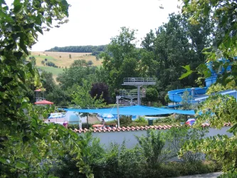 Ein Schwimmbad mit blauen Rutschen und Liegen in einer grünen Umgebung. Im Hintergrund sind sanfte Hügel zu sehen.