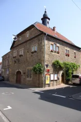 Ein historisches Gebäude aus Stein an einer Straßenecke. Es hat ein rotes Dach, Blumenkästen und einen kleinen Turm.