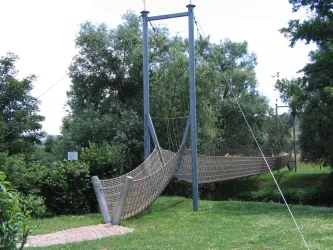 Eine Hängebrücke aus Seil und Metall überspannt einen kleinen Graben. Umgeben ist sie von grünen Bäumen und einer Wiese.