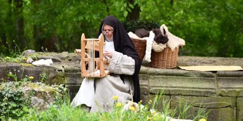 Eine Nonne sitzt im Freien und arbeitet an einer Holzarbeit. Im Hintergrund sind Körbe mit Wolle und anderen Materialien zu sehen.