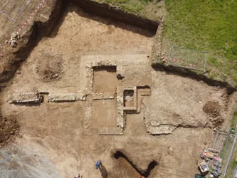 Eine archäologische Ausgrabungsstelle mit gut erkennbaren Fundamenten und Gräben. Grüne Wiese umgibt den Bereich, während Werkzeuge und Materialien in der Nähe liegen.