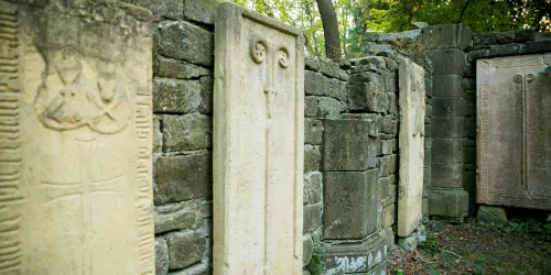 Eine Reihe von alten Steintafeln stehen in einem bewaldeten Bereich. Die Tafeln sind mit verschiedenen reliefartigen Mustern verziert.