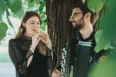 Ein Paar spielt Musik unter einem Baum. Die Frau spielt eine Flöte, während der Mann ein Akkordeon spielt.
