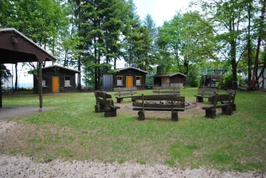 Ein ruhiger Platz mit Holzliegen und einem Lagerfeuerkreis. Im Hintergrund sind kleine Holzferienhäuser und zahlreiche Bäume zu sehen.