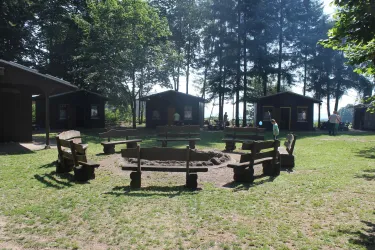 Ein gemütlicher Lagerfeuerplatz mit Holzbänken und einem zentralen Feuerkreis. Im Hintergrund sind einige Hütten und Bäume zu sehen.