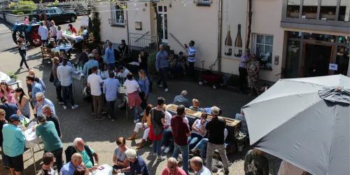 Eine lebhafte Veranstaltung im Freien mit vielen Menschen, die an Tischen sitzen und miteinander sprechen. Die Umgebung zeigt ein geselliges Ambiente mit Musik und Essensständen.