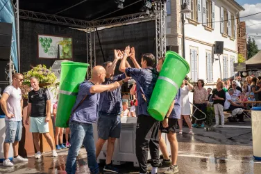 Ein fröhliches Fest mit Menschen, die in blauen Shirts und grünen Behältern um die Wette tanzen. Im Hintergrund sind Zuschauer und eine festliche Atmosphäre zu sehen.