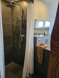 Eine moderne Dusche mit dunkelgrauen Fliesen und einem transparenten Duschvorhang. Die Wände sind hell, mit einem Holzregal für Pflegeprodukte.