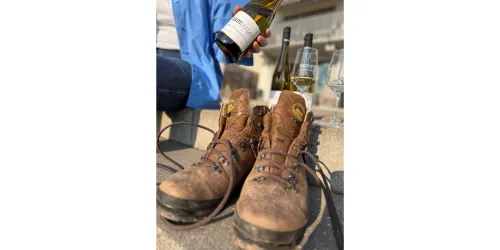 Ein Paar braune Wanderschuhe stehen auf einer Mauer. Im Hintergrund wird eine Flasche Wein gehalten, und eine Glas Wein steht daneben.