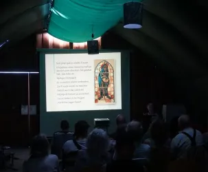 Eine Präsentation in einem rustikalen Raum mit einer Gruppe von Zuhörern. Auf der Leinwand ist ein Bild eines bunte Glasfensters mit Text zu sehen.