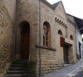 Ein historisches Gebäude aus Sandstein mit großen Fenstern und einer charakteristischen Fassade. Die Eingangstür ist aus Holz und die Straße ist gepflastert.