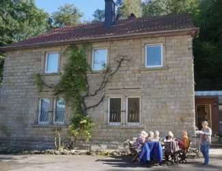 Ein Steinhaus umgeben von Bäumen. Eine Gruppe von Personen sitzt entspannt an einem Tisch draußen.