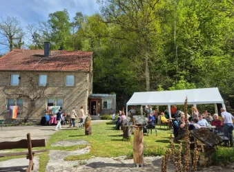 Ein schönes Haus inmitten von Bäumen mit einer bunten Gruppe von Menschen im Freien. Im Vordergrund gibt es Tische und eine Zeltüberdachung für eine Veranstaltung.