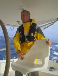 Ein Mann in einer gelben Segeljacke steht auf einem Boot. Er hält einen Stuhl und blickt konzentriert in die Ferne.
