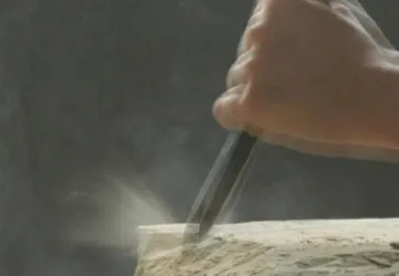 Eine Hand bearbeitet einen Stein mit einem Meißel. Staub wird aufgewirbelt, während der Stein bearbeitet wird.