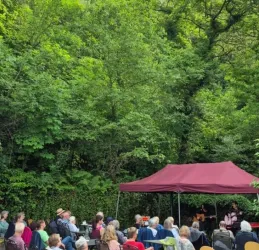 Eine Gruppe von Menschen sitzt unter einem Zelt in einem grünen Wald. Auf der kleinen Bühne treten Musiker auf.
