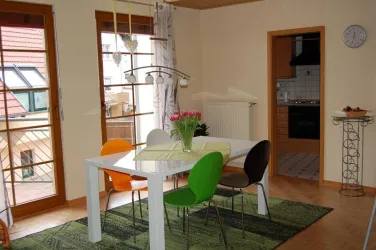 Ein helles Esszimmer mit einem weißen Tisch und bunten Stühlen in grünen Tönen. Auf dem Tisch steht eine Vase mit frischen Blumen, und die Fenster lassen viel Licht herein.