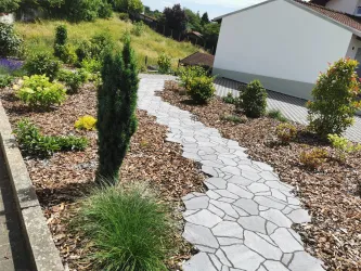 Ein schöner Gartenweg aus Steinen führt durch eine abwechslungsreiche Pflanzenlandschaft. Verschiedene Bäume und Sträucher umgeben den Weg und verleihen dem Bereich eine freundliche Atmosphäre.