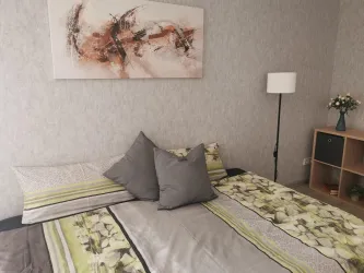 Ein gemütliches Schlafzimmer mit einem Bett, das mit grünen und grauen Kissen dekoriert ist. Im Hintergrund sind eine Lampe und ein Regal mit einer Blumendekoration zu sehen.