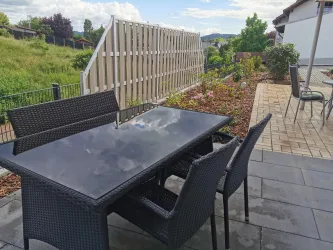 Ein moderner Esstisch aus Rattan mit Stühlen auf einer Terrasse. Im Hintergrund sind gepflegte Pflanzen und ein Sichtschutz zu sehen.