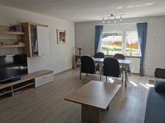 Ein modernes Wohnzimmer mit einem großen Fenster und natürlichem Licht. Es gibt einen Fernseher, einen Esstisch und stilvolle Möbel.