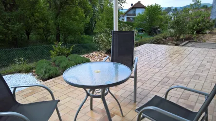 Eine gemütliche Terrasse mit einem runden Tisch und zwei Stühlen. Im Hintergrund sieht man einen grünen Garten mit Bäumen und Sträuchern.