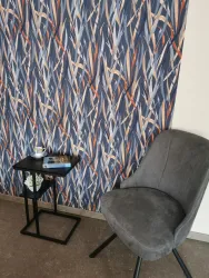 Ein gemütlicher Sessel steht neben einem kleinen Tisch mit einer Tasse und einem Buch. Im Hintergrund ist eine farbenfrohe Tapete mit einem abstrakten Muster zu sehen.