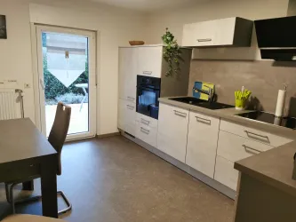 Eine moderne Küche mit hellen Schränken und einem Tisch. Durch ein Fenster ist ein Blick auf einen Außenbereich sichtbar.