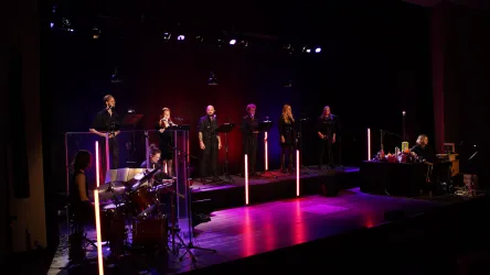 Eine musikalische Darbietung auf einer Bühne mit mehreren Sängern und einem Schlagzeuger. Die Bühne ist mit bunten Lichtern beleuchtet und es gibt transparente Trennwände.