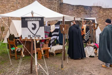 Ein mittelalterlicher Marktstand mit historischen Kleidern und Utensilien. Besucher in Gewändern erkunden die Stände unter einem Zelt.