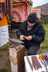 Ein Handwerker arbeitet an einem Tisch mit verschiedenen Lederriemen und Werkzeugen. Im Hintergrund ist ein Zelt mit dekorativen Elementen zu sehen.