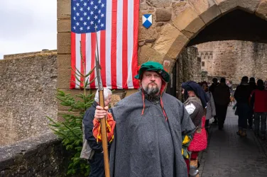 Ein Mann in historischem Gewand steht vor einem Tor, während er einen Stock hält. Im Hintergrund ist eine amerikanische Flagge und eine Menschenmenge zu sehen.