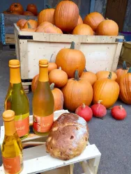 Eine Vielzahl von orangefarbenen Kürbissen liegt in einem Holzbehälter. Darauf stehen Flaschen mit Getränken und ein Laib Brot.