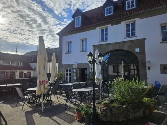 Ein ruhiger Innenhof mit Tischen und Stühlen vor einem Gebäude. Sonnenschirme und Pflanzen schaffen eine einladende Atmosphäre.