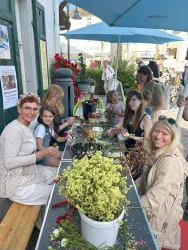 Eine Gruppe von Menschen sitzt an einem Tisch und bastelt mit Blumen. Es ist eine freundliche Atmosphäre, und die Teilnehmer scheinen Freude an der Aktivität zu haben.