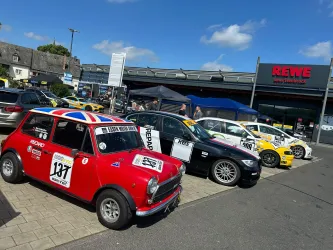 Eine Reihe von Autos steht auf einem Parkplatz vor einem Supermarkt. Das auffälligste Auto ist ein roter Mini mit einer Union-Jack-Flagge auf dem Dach.
