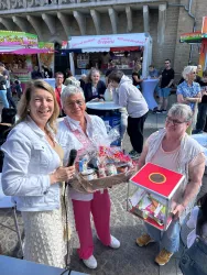 Eine fröhliche Veranstaltung im Freien mit mehreren Personen. Eine Frau präsentiert stolz einen Geschenkkorb und wird von zwei anderen unterstützt.