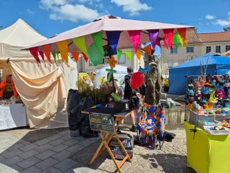 Ein lebhafter Marktstand mit bunten Fahnen und Dekorationen. Menschen schlendern vorbei und genießen die fröhliche Atmosphäre.