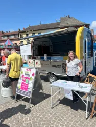 Ein Verkaufsstand auf einem Marktplatz mit einem bunten Verkaufswagen. Eine Frau steht lächelnd daneben und es gibt verschiedene Informationsmaterialien auf Tischen.
