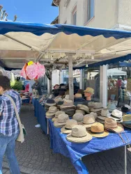 Ein Marktstand mit vielen verschiedenen Hüten steht unter einem blauen Zelt. Im Hintergrund sind Menschen zu sehen, die sich umschauen.