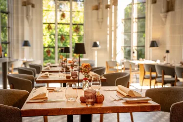 Ein elegantes Restaurant mit Holztischen und stilvollen Stühlen. Große Fenster lassen viel Licht herein und bieten einen Blick auf den Garten.