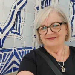 Eine Frau mit grauen Haaren und Brille lächelt in die Kamera. Im Hintergrund sind blaue Fliesenmuster zu sehen.